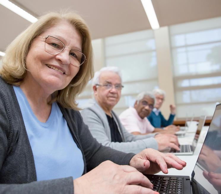 zdjęcie 4 uśmiechniętych seniorów w rzędzie siedzą przy stołach przy laptopach 