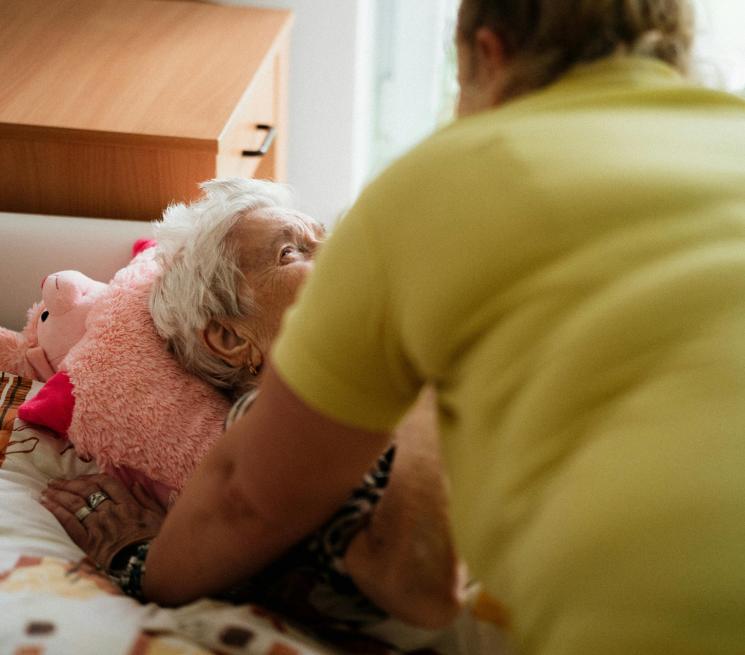 zdjęcie opiekunka opiera się o łóżko leżącej podopiecznej i z nią rozmawia