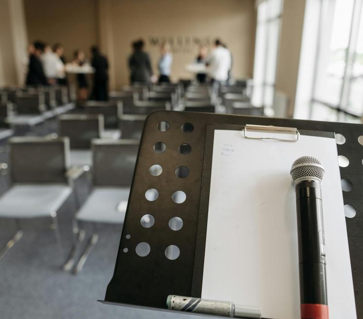zdjęcie sala konferencyjna na pierwszym planie mównica na niej leży biała kartka oraz mikrofon w tle kilka osób 