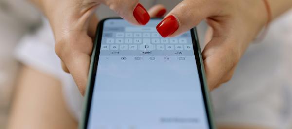 zdjęcie  dłonie kobiety z czerwonymi paznokciami, kótra trzyma smartfona i pisze wiadomość tekstową