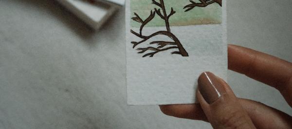 zdjęcie kobieta trzyam w dłoni zakładkę do książki na niej rysunek pałacu gałęzie i liście drzew w tle leżą książki