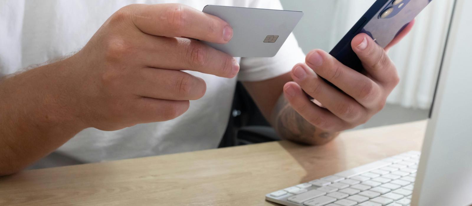 zdjęcie dłonie trzymające kartę bakomatową i telefon na biurku leży laptop